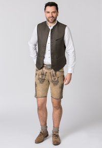 Traditionelles Herrenoutfit mit einer strukturierten grünen Weste, einem weißen Hemd, beigen Lederhosen mit gestickten Designs und braunen Schuhen mit Socken.