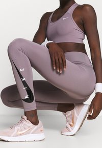 Leggings de sport violets avec le logo Nike noir, associés à un soutien-gorge de sport violet assorti et des baskets Nike rose clair. Matière douce et extensible.