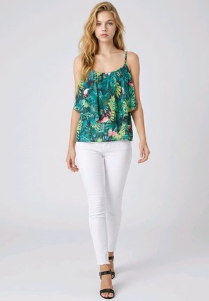 Joven mujer con una camiseta sin mangas verde con estampado tropical, vaqueros pitillo blancos y sandalias negras con tiras en el tobillo, de pie frente a un fondo sencillo.