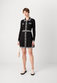 Robe noire avec des accents blancs, présentant des manches longues, un col et des boutons sur le devant. Associée à des mocassins de couleur claire et un sac.