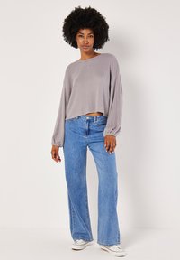 Top cropped grigio a coste con maniche lunghe abbinato a jeans blu a gamba larga e sneaker bianche. Il modello è in piedi su uno sfondo chiaro.