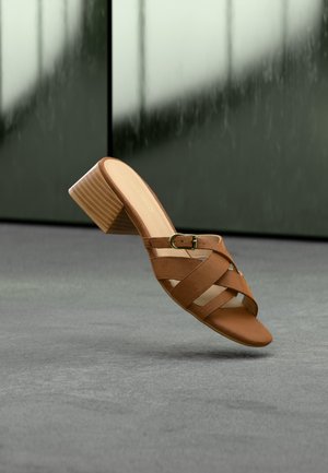 Mule en cuir marron à bout ouvert, avec un talon bloc et des brides croisées, posé sur un sol gris avec un arrière-plan sombre flou.