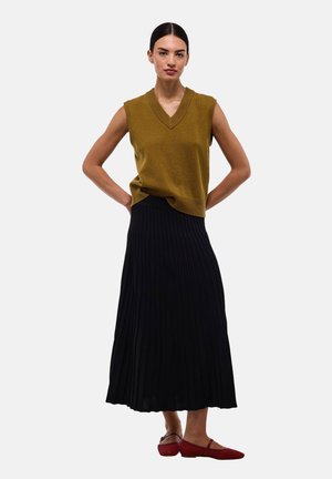 Femme debout, mains sur les hanches, portant un haut en maille marron sans manches, une jupe midi plissée noire et des chaussures plates rouges.