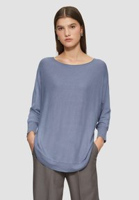 Helles blaues, locker sitzendes Sweatshirt mit langen Ärmeln und weitem Ausschnitt, das eine weiche Textur und einen abgerundeten Saum aufweist. Über grauen Hosen getragen.