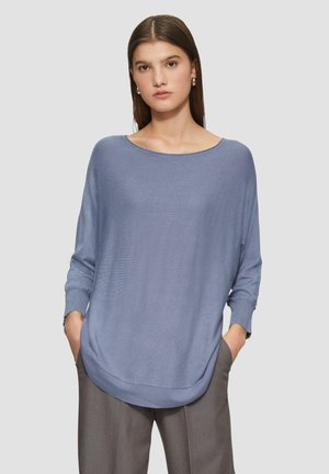 Helles blaues, locker sitzendes Sweatshirt mit langen Ärmeln und weitem Ausschnitt, das eine weiche Textur und einen abgerundeten Saum aufweist. Über grauen Hosen getragen.