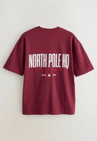 Bordová bavlnená tričko s krátkymi rukávmi. Obsahuje biely potlač: "NORTH POLE HQ," "25 YULE ROAD," a "ESTD 1925" s ikonou stromu a akcentami.