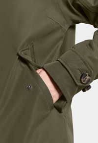 Olijfgroene regenjas met een knoopsluiting bij de manchetten, een borstzak en zijzak met zichtbare stiksels en hardwaredetails.