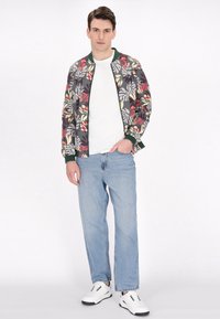 Bloemige bomberjack in grijs, rood en geel, met een groene rand. Gepaard met lichtblauwe jeans en witte sneakers. Eenvoudig wit T-shirt eronder.