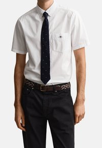 Homme portant une chemise blanche à manches courtes avec poche avant, cravate bleu marine à pois, ceinture tressée marron et pantalon noir, debout devant un fond blanc.