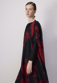 Garbaggio nero con un design fluido, maniche lunghe e una silhouette simile a un mantello. Decorato con grandi motivi floreali rossi.