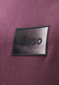 Maglietta bordeaux con una texture liscia che presenta un'etichetta logo rettangolare nera con la scritta "HUGO" in rilievo in caratteri bold.