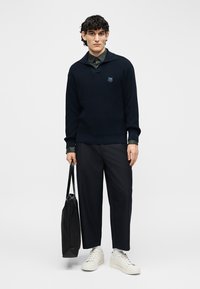 Νέος άνδρας στέκεται κρατώντας μια μαύρη τσάντα ώμου, φορώντας μαύρο πουλόβερ, σκούρο παντελόνι και λευκά αθλητικά παπούτσια, μπροστά σε ένα απλό φωτεινό φόντο.