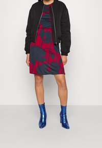 Veste bomber noire sur une robe à motifs floraux bleus et rouges à manches courtes. Bottines brillantes bleues. Fond simple.