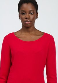 Maglione rosso lavorato a maglia con un ampio scollo e maniche lunghe. Il tessuto sembra morbido e strutturato, con una silhouette aderente su uno sfondo chiaro.