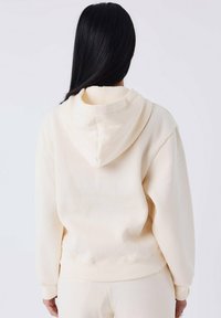Persona con lunghi capelli neri che indossa una felpa beige chiaro e pantaloni abbinati, mostrata di spalle su uno sfondo neutro.
