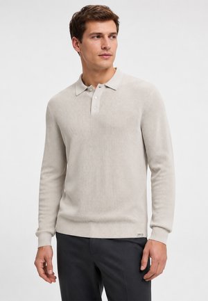 Man draagt een beige poloshirt met lange mouwen en donkere broek, kijkt iets naar rechts tegen een effen achtergrond.