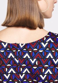 Robe multicolore présentant des motifs géométriques rouges, bleus et blancs sur un fond noir, avec une fermeture éclair discrète dans le dos et un large décolleté.