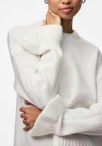 Λευκό πλεκτό πουλόβερ με ριμπ μανσέτες και στρογγυλή λαιμόκοψη, με απαλή υφή και άνετη εφαρμογή. Το μοντέλο έχει τα χέρια σταυρωμένα χαλαρά.