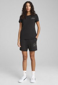 Must mustrepp ja lühikesed püksid, millel on broderi "Puma" logo. Kompositsioon sisaldab valgeid tossusid ja ribatud pahkluusokke, luues sportliku ilme.