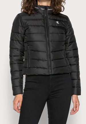 Kvinde iført sort Calvin Klein pufferjakke med lynlås og sidelommer, kombineret med sorte bukser, mod en ensfarvet baggrund.