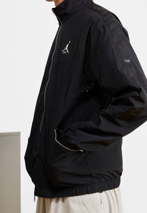 Person trägt eine schwarze Jacke mit Reißverschluss und einem weißen Jumpman-Logo auf der Brust, die Hände in den seitlichen Taschen, kombiniert mit beigen Hosen.