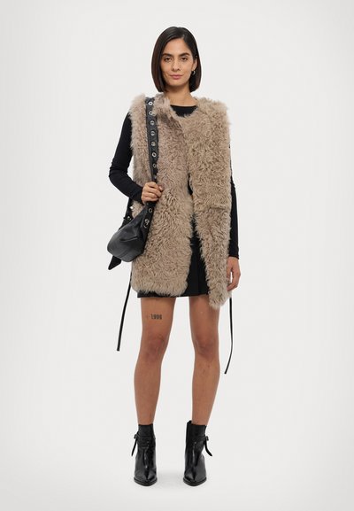 Neppelsen vest in beige, met een textuurafwerking, open voorkant, gedragen over een zwart langemouwen top en minirok. Zwarte handtas met schouderband.