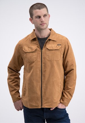 Beige zip-up jas met een gladde textuur, voorzien van twee borstzakken, een kraag en lange mouwen. Gedragen over een zwart shirt.