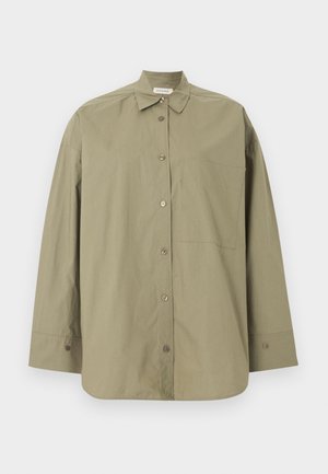 Γυναίκα στέκεται φορώντας υπερμεγέθη ελιά πουκάμισο, λευκό παντελόνι με τελειώματα με τρύπες, καφέ σανδάλια, κρατώντας μεγάλη μαύρη τσάντα σε απλό φόντο.