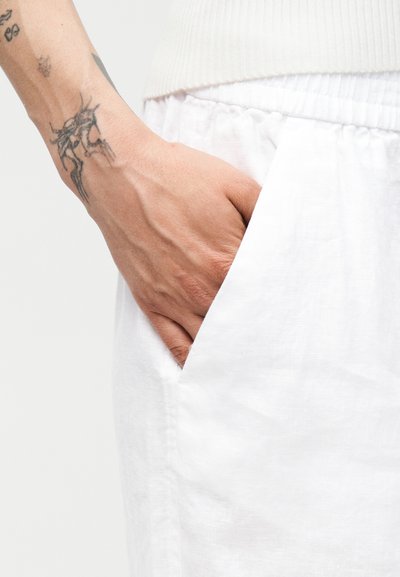 Main droite avec des tatouages insérée dans la poche d'un pantalon blanc à taille élastique, partie visible d'un haut blanc côtelé au-dessus.
