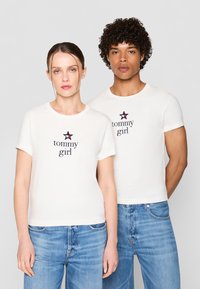 Hvite bomull T-skjorter med korte ermer som har en svart stjerne og "tommy girl" tekst i marineblått. Brukt med blå denimjeans.