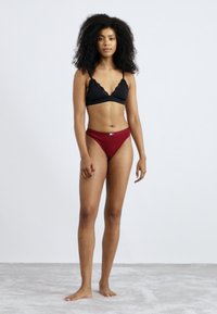 Zwarte scalloped bralette en burgundy thong. De bralette heeft dunne bandjes en een kantafwerking; de thong heeft een klein merketiket.