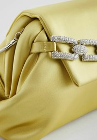 Kollane satiinkotike, millel on tekstuuriga pind, kaunistatud rhinestone-i klapiga ja sujuva, ümarate joontega disain.