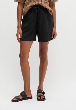 Persoon draagt zwarte shorts met trekkoord, een bruin gebreide top en zwarte slippers met een gouden gesp, staand tegen een effen achtergrond.