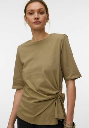 Femme portant une chemise kaki à manches courtes avec un nœud latéral, des boucles d'oreilles en perles et un bracelet à motif zèbre à son poignet gauche.