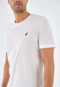 White short-sleeve t-shirt made of soft fabric, featuring a round neckline and a small black bird logo on the left chest.