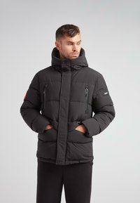 Mand i sort quilted puffer jakke med høj krave, lynlåslommer på brystet, sidelommer og logo-patch på den øverste ærme, hænderne i lommerne.
