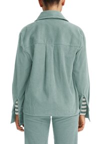 Camicia in corduroy verde menta con colletto, dettaglio della piega sul retro e spacchi laterali. Presenta un tessuto a righe che fa capolino dai polsini.