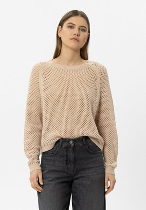 Woman wearing a beige knit sweater and high-waisted black jeans, standing against a plain white background, facing forward with a neutral expression.