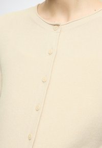 Light beige cardigan with a round neckline, featuring six matching buttons down the front and a soft, textured knit fabric.