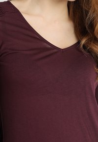 Burgundy long-sleeve top with a V-neckline, made of soft material. Features lace accents on the shoulders. Smooth texture, fitted design.