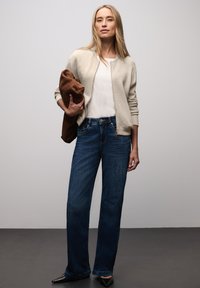 Frau in einem beigen Reißverschluss-Pullover, weißem Oberteil, blauer Jeans und schwarzen Schuhen, die eine gefaltete braune Jacke vor einem grauen Hintergrund hält.