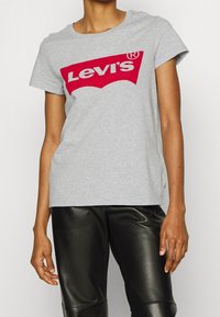 Šedé bavlněné tričko s výrazným červeným logem Levi's na přední straně, krátké rukávy a volný střih, spárované s černými koženými kalhotami.