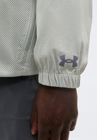 Μανίκι ανοιχτόγκρι υφασμάτινου σακακιού με ελαστικό μανσέτα και σκούρο λογότυπο Under Armour πάνω από τον καρπό σε σκουρόχρωμο δέρμα.