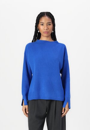 Pull en tricot bleu avec une texture côtelée, doté d'une coupe décontractée, de manches longues et d'entonnoirs sur les côtés à l'ourlet. Associé à un pantalon sombre.