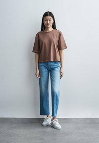 Žena stojaca pri obyčajnej stene, oblečená v hnedej oversize tričku, svetlomodrých kraťasových džínsoch a bielych teniskách.