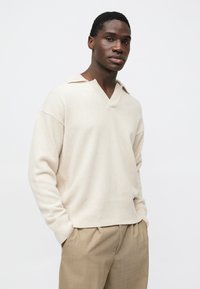 Pull en maille beige avec un col en V et un petit col. Texture douce, coupe ample et poignets côtelés. Associé à un pantalon plissé marron clair.