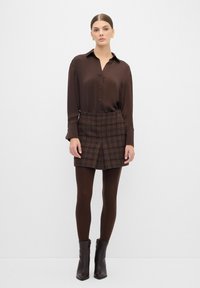 Blusa di seta marrone con bottoni, abbinata a una mini gonna a quadri marrone. Il modello indossa collant marroni e stivaletti. Completo semplice ed elegante.