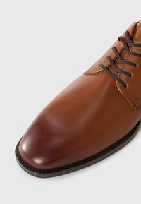 Zapato de vestir de cuero marrón con cordones y un acabado degradado que va de un tono claro de tinte tostado a un burdeos profundo, punta redondeada y suela negra con detalle de costura.