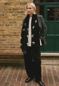 Blond kvinde står op ad en murstensvæg iført et sort tørklæde med hvide sløjfer, grå blazer, hvid sweater, sorte bukser og brune sko.