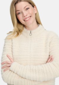 Fuzzy kremowy sweter z wysokim kołnierzem, z zamkiem błyskawicznym z przodu i strukturalnym ryżowym splotem. Długie rękawy z dopasowanymi mankietami.